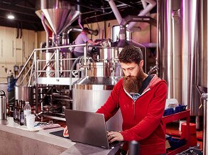 A bearded man uses a laptop in a craft brewery, managing operations and cash flow for his food and beverage business.