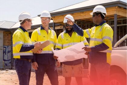 Four construction workers in hard hats and high-vis vests discuss plans on a building site, highlighting the need for consistent working capital.