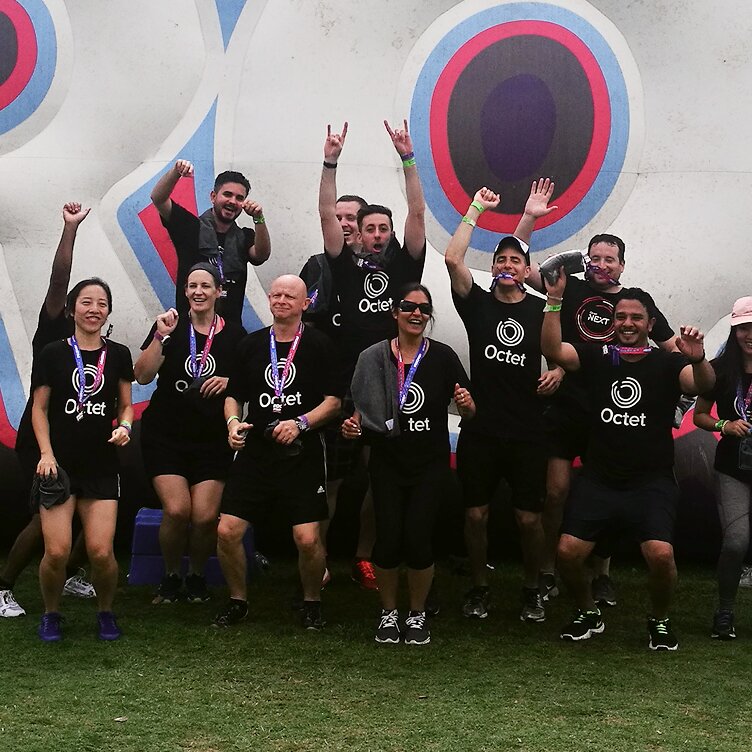A diverse group of Octet employees in black t-shirts cheer enthusiastically at an outdoor team event.